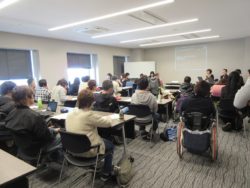 Photo taken from the back of the seminar room. The audience is seen from behind, sitting in rows. Julia and You are sitting behind a table at the front, facing the audience. To their left on the wall is Julia’s power point presentation in Japanese