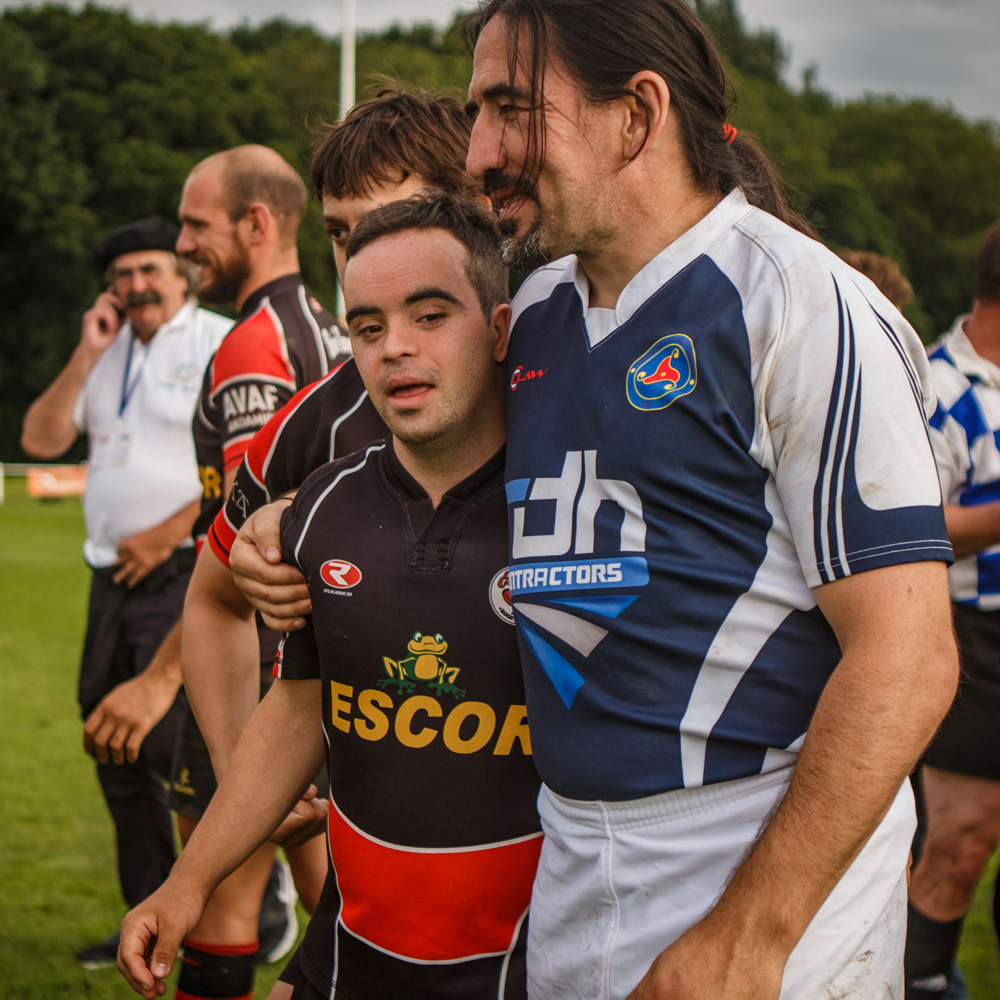 Two Rugby players after a match, team mates in background. One player is non-disabled, the other is a disabled person who has Downs Syndrome.
