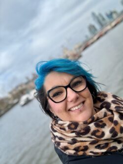 Photograph of Johanna Knebel. She has black hair with an electric blue streak at the front, black glasses and a lip ring. She is smiling and standing in front of a city river